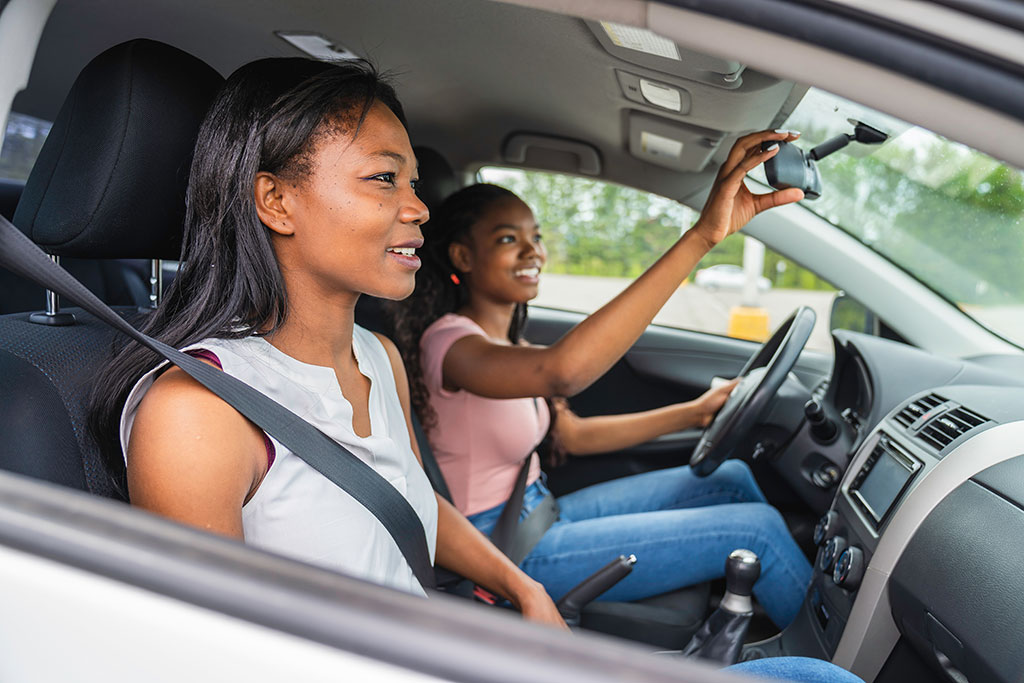 teen behind the wheel of a car adjusting rear view mirror with parent in passenger seat.